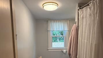 A bathroom with a toilet, a window with a view, a shower curtain, and a basket of toiletries.