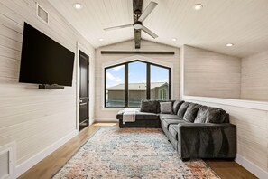 A room with a large window, a ceiling fan, a flat-screen TV, and a gray sofa with multiple cushions. - Luxe 3BR Lake Stay | BBQ | Firepit | Pickleball (Spicewood)
