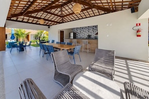 An outdoor dining area with a wooden table and chairs, a kitchen in the background, and a fire extinguisher on the wall. - Apt. w/ Balcony in Condo. w/ Pool Pim0083 (Camaçari)