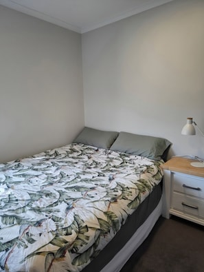 A single bed with a floral bedspread, a bedside table with a lamp, and a white wall. - 2-minutes walk to the beach, Spacious Home and great outdoor living. (Tauranga)