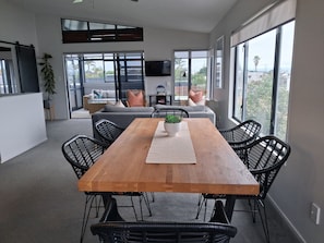 A modern living room with a wooden dining table, black wicker chairs, a grey sofa, and a large window offering a view of the outdoors. - 2-minutes walk to the beach, Spacious Home and great outdoor living. (Tauranga)