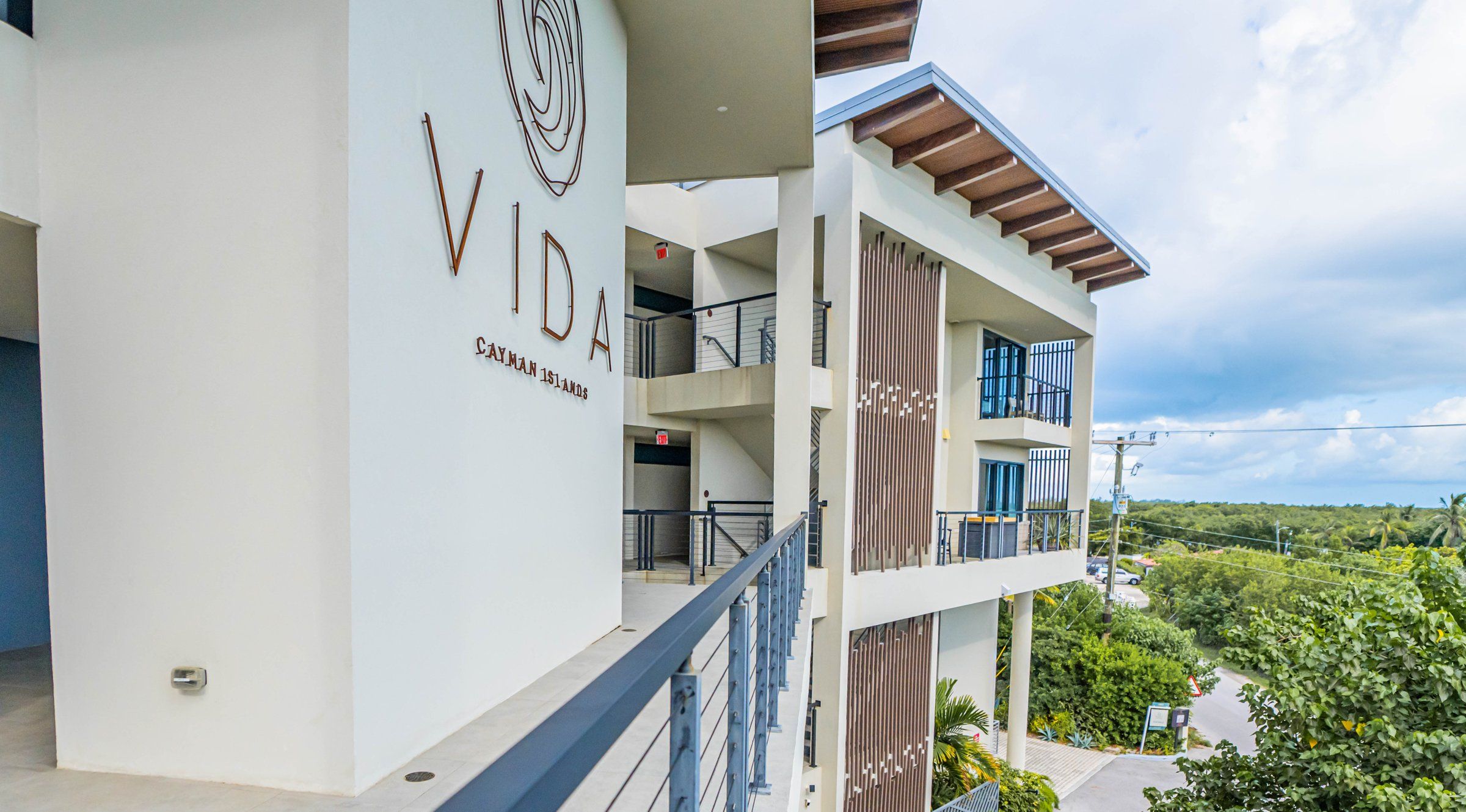 Un edificio de varios pisos con un balcón, un cartel que dice 'VIDA Cayman Islands' y vistas a zonas verdes y una carretera.