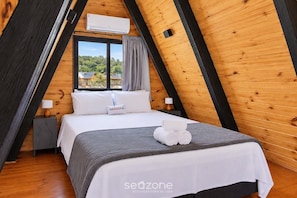 A bedroom with a sloped ceiling, a bed with white bedding, a window with curtains, and an air conditioning unit. - Cabin With hot tub in Condo. With Pool (Pirenópolis)