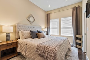 A bedroom with a large bed, a nightstand, a lamp, and a wall-mounted clock. - Southern Row | Two King Bedrooms | Walk to UA (Tuscaloosa)