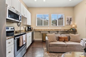 A compact kitchen with a microwave, stove, and oven, a beige sofa with striped pillows, and a window with blinds. - Southern Row | Two King Bedrooms | Walk to UA (Tuscaloosa)