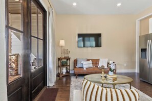 A living room with a brown sofa, a coffee table, and a television on the wall. - Southern Row | Two King Bedrooms | Walk to UA (Tuscaloosa)