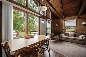 A spacious room with a wooden dining table, chairs, and a sofa. Large windows provide views of the outdoors. - Seehaus at Sommer Dorf (West Branch)