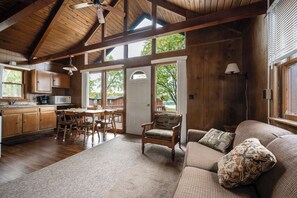 A spacious room with a wooden ceiling, a dining area with a table and chairs, a sofa with patterned cushions, and a view of the outdoors through large windows. - Seehaus at Sommer Dorf (West Branch)