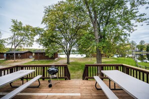 A wooden deck with picnic tables and a barbecue grill, overlooking a lake with cabins in the background. - Seehaus at Sommer Dorf (West Branch)