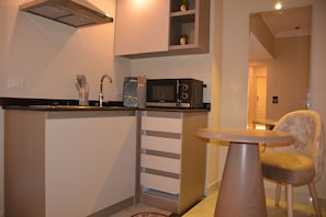 Cocina compacta con microondas, un lavamanos y un mueble pequeño. Hay una silla y una mesa en primer plano.