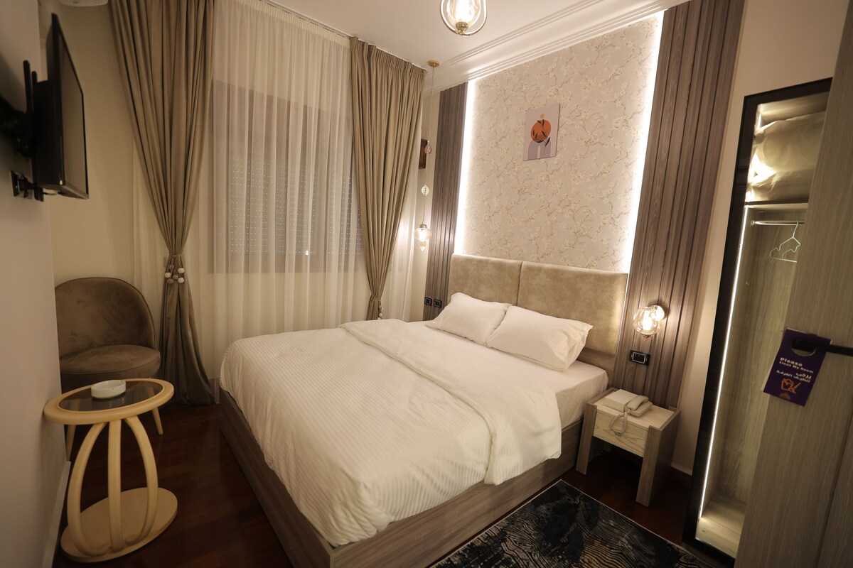 Habitación de hotel con una cama grande, una mesita de noche con lámpara, una silla y un televisor montado en la pared.