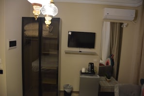 Una habitación de hotel con cama, televisor, escritorio con silla, un cesto de basura y un equipo de aire acondicionado.