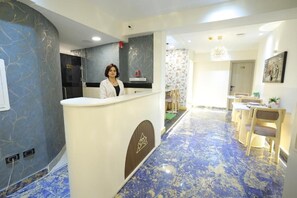 Zona de recepción de un hotel, con recepcionista, mostrador de recepción, comedor con mesas y sillas, y piso estampado.