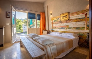 A bedroom with a large bed, a bench, a wooden headboard with a painting, and a view of the outdoors through a glass door. - Amazing home in Serra San Quirico with outdoor swimming pool (Serra San Quirico)