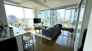 A modern living room with a sofa, a dining area with a table and chairs, and large windows offering a city view.