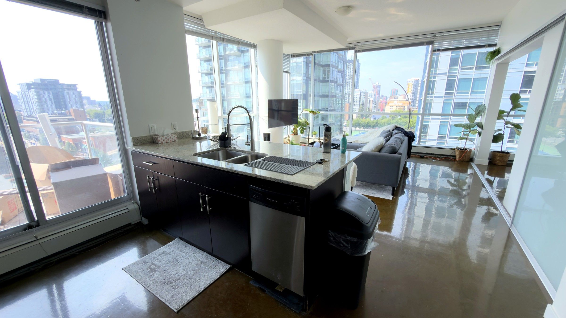 A modern kitchen with a large window, a built-in dishwasher, and a grey sofa in the living area.