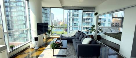 A modern living room with a sofa, coffee table, and a TV. Large windows offer a view of the cityscape.