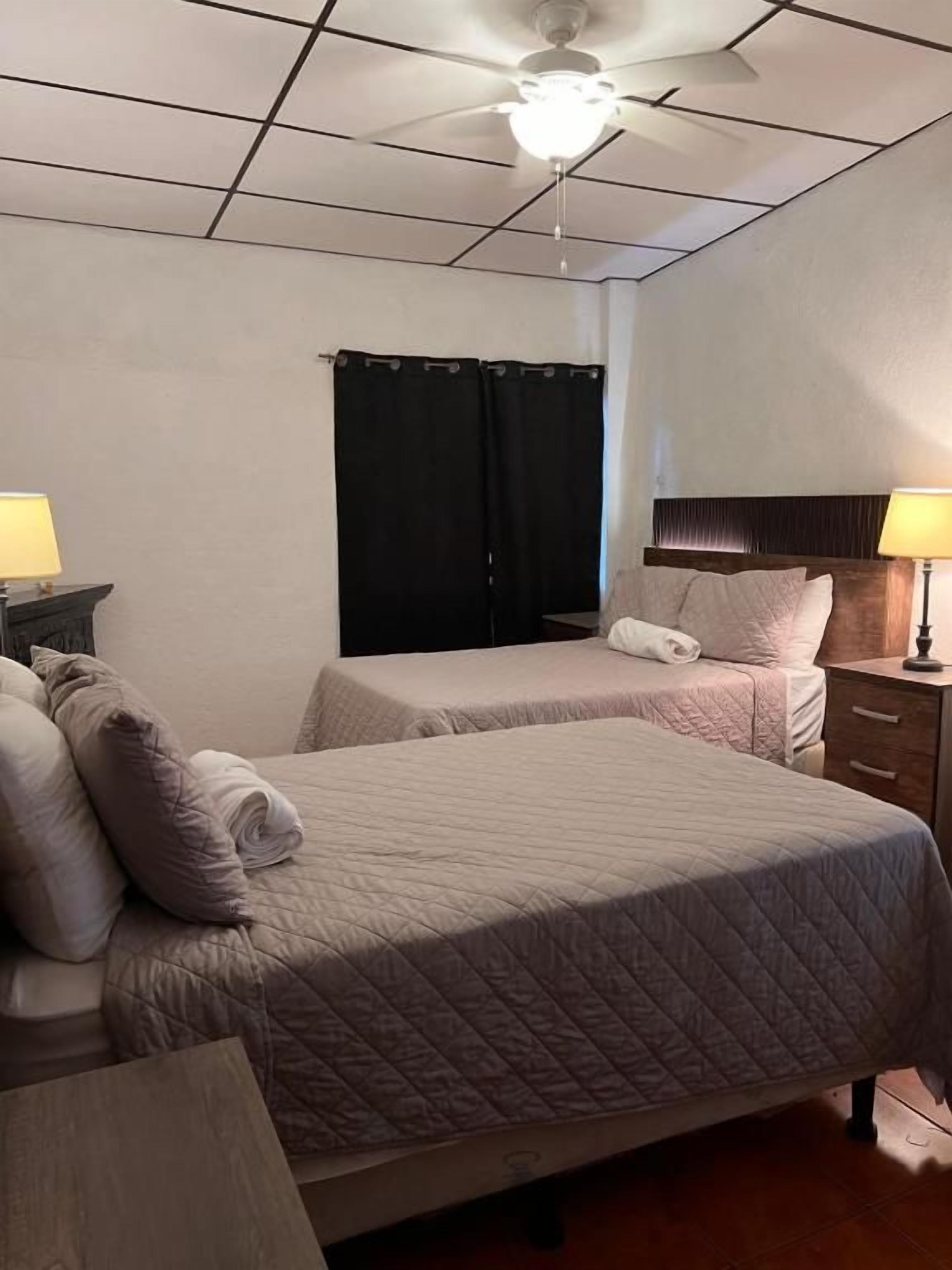 Habitación de hotel con dos camas, ventilador de techo, ventana con cortinas y lámparas de noche.