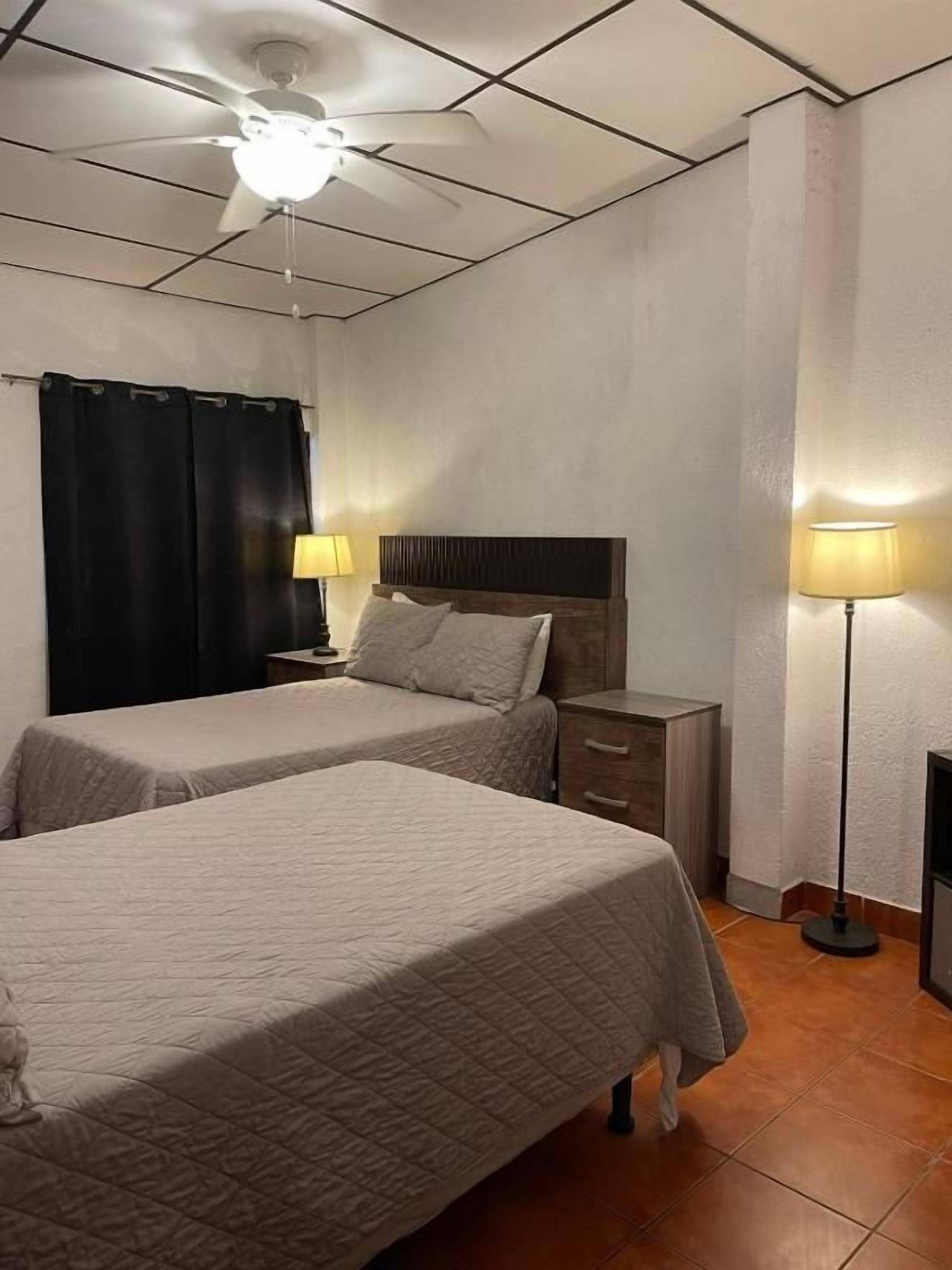Ett hotellrum med två sängar, en takfläkt, ett nattduksbord med en lampa och en väggmonterad TV.