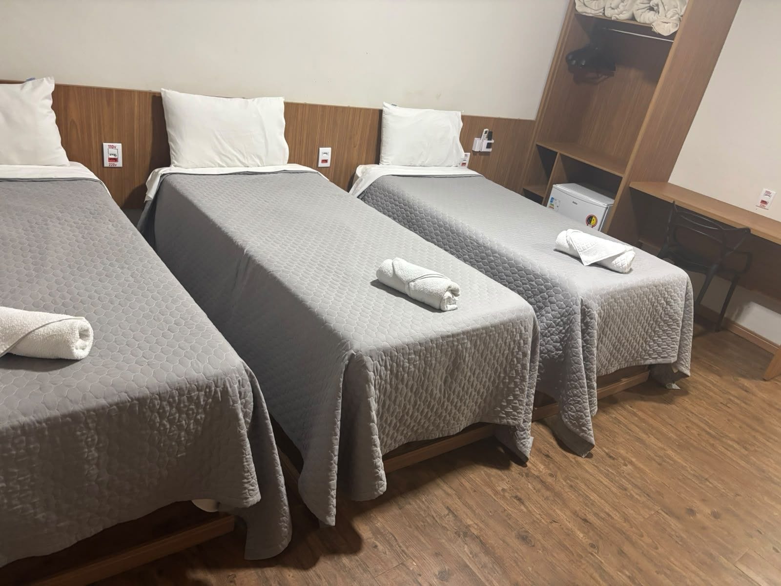 Een hotelkamer met drie eenpersoonsbedden, elk voorzien van een grijs gewatteerde spreidek en witte kussens. Er is een houten garderobe en een klein tafeltje met een stoel.