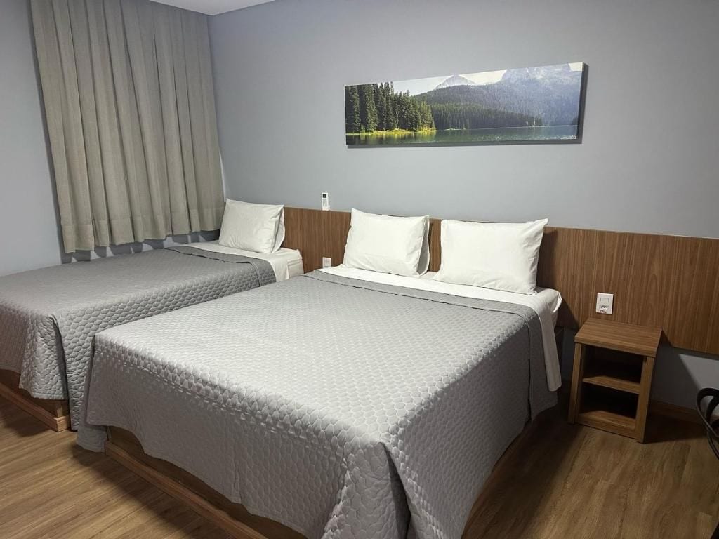 Habitación de hotel con dos camas, un cabecero de madera, una mesita de noche y un cuadro de un lago y montañas en la pared.