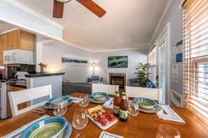 A dining area with a wooden table set for four, surrounded by chairs. There is a fireplace, a painting on the wall, and a kitchen in the background. - North Mission Beach House - Patio + Garage (San Diego)