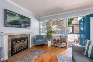 A living room with a fireplace, a blue armchair, a wooden coffee table, a bookshelf with board games, and a view through a window with blinds. - North Mission Beach House - Patio + Garage (San Diego)