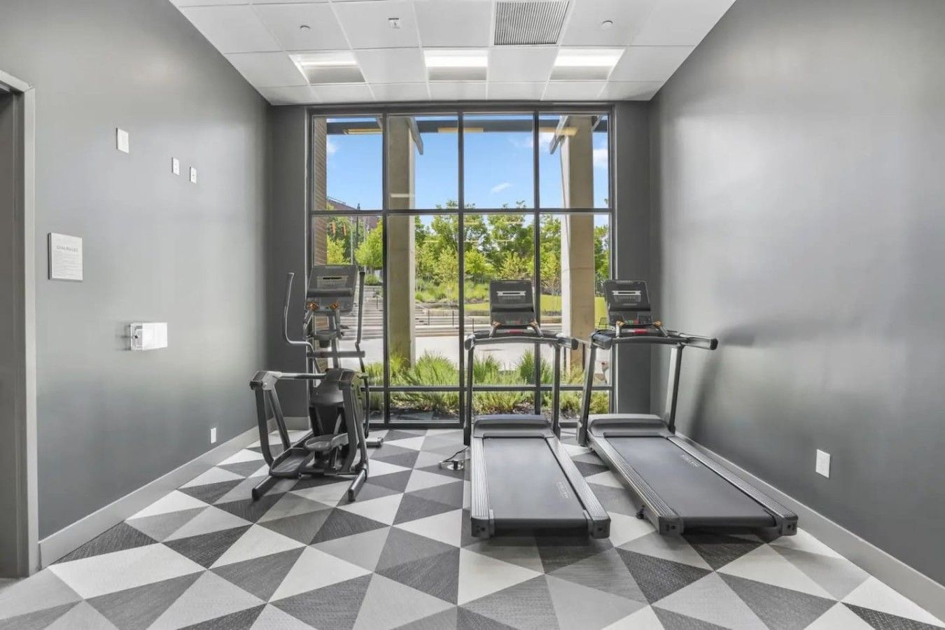 A gym with treadmills, an elliptical, and a stationary bike, with large windows and a geometric patterned floor.