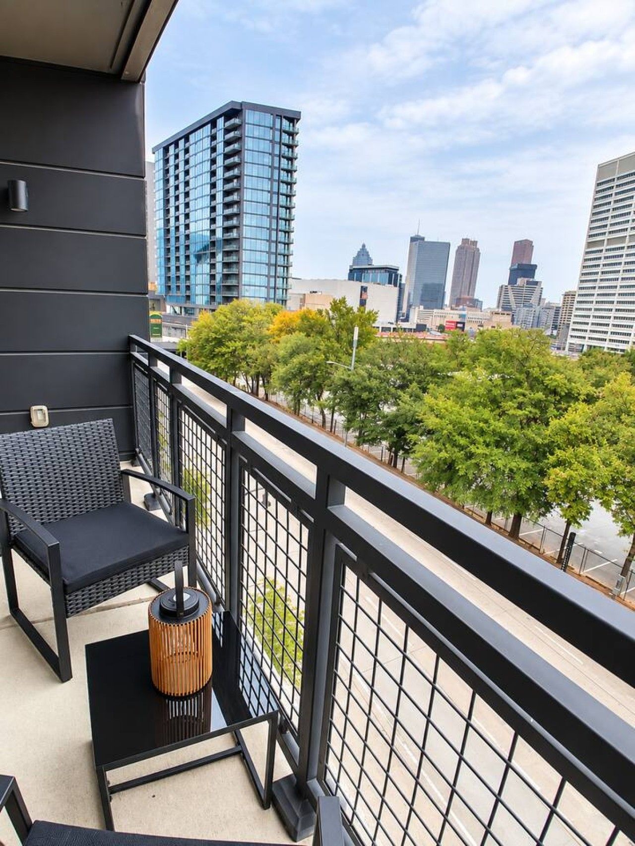 A balcony with a black metal railing, a black sofa, and a small table with a cylindrical lamp. In the background, there are tall buildings and trees.