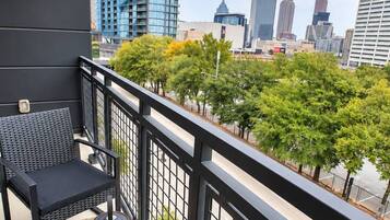 A balcony with a black metal railing, a black sofa, and a small table with a cylindrical lamp. In the background, there are tall buildings and trees.