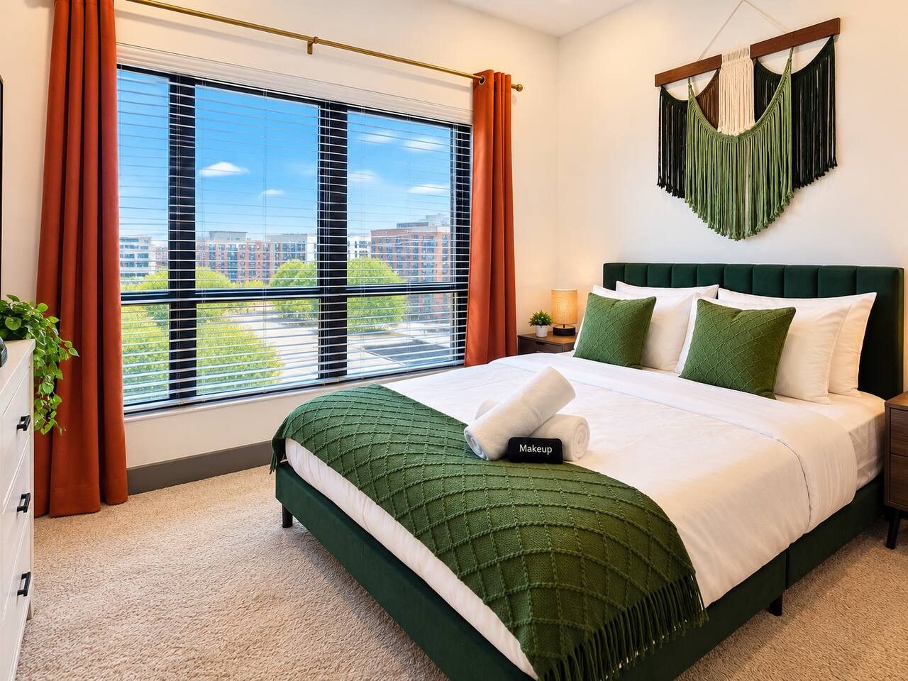 A hotel room with a large window, a bed with green bedding, a nightstand, and a hanging macrame wall decoration.