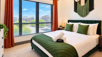 A hotel room with a large window, a bed with green bedding, a nightstand, and a hanging macrame wall decoration.