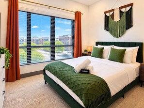 A hotel room with a large window, a bed with green bedding, a nightstand, and a hanging macrame wall decoration. - Stadium/Events Suite Spot I Gym & Rooftop Lounge (Atlanta)