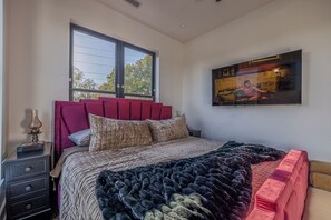 Quarto com cama, mesa de cabeceira, janela e televisão fixada na parede.