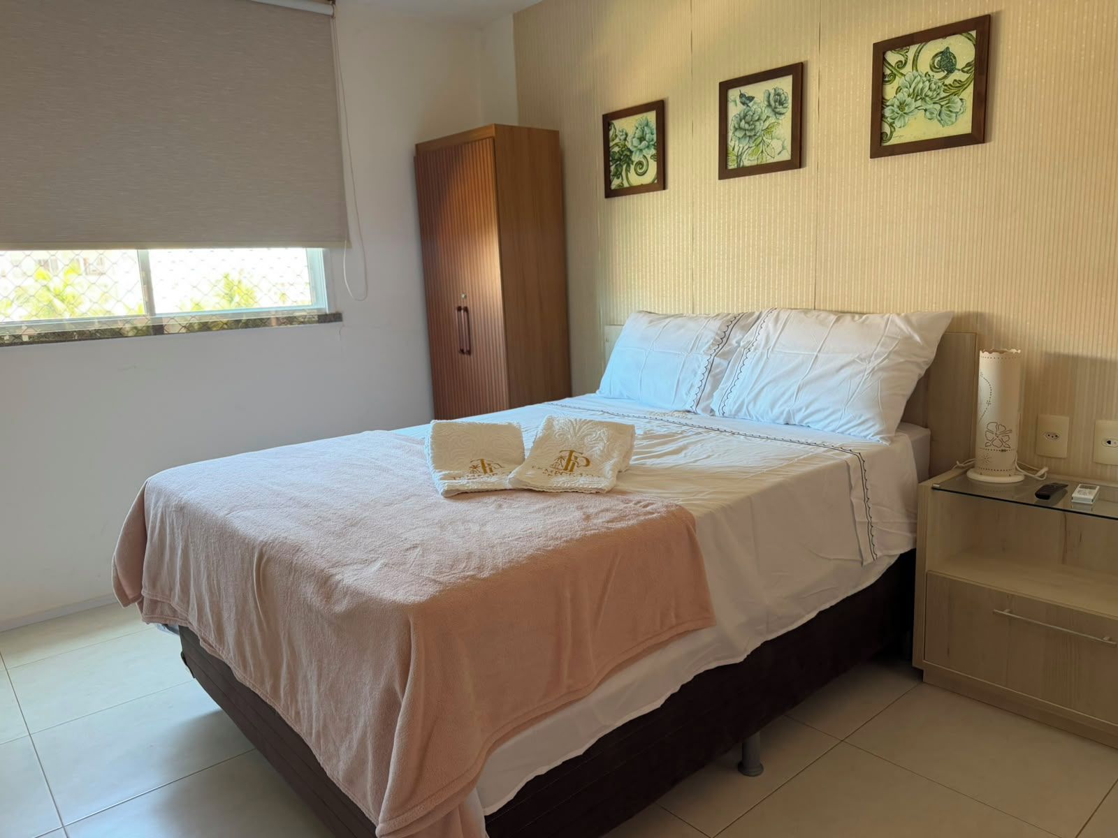 Una cama bien hecha con una colcha blanca y color melocotón, una mesita de noche con una lámpara, y una ventana con vistas al follaje.