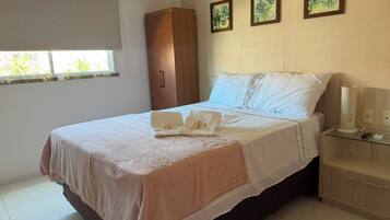 Una cama bien hecha con una colcha blanca y color melocotón, una mesita de noche con una lámpara, y una ventana con vistas al follaje.