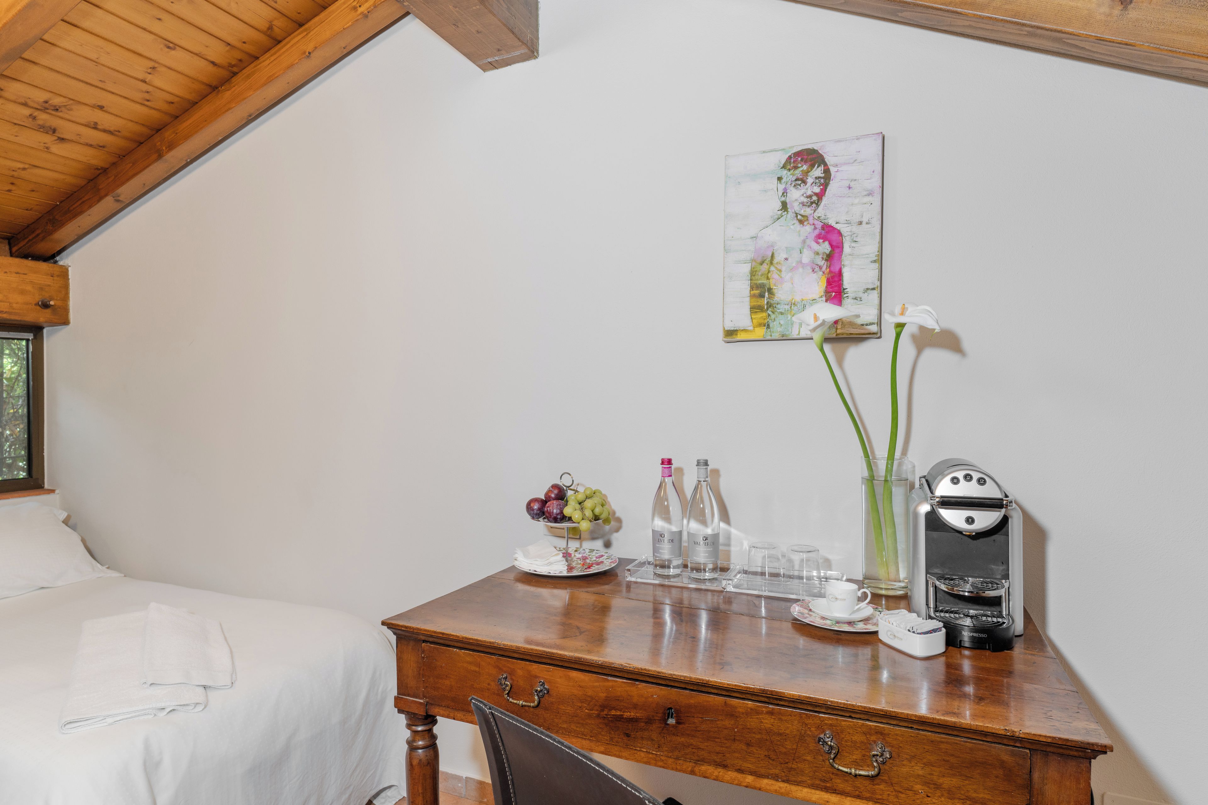 A wooden desk with a coffee maker, two bottles, a vase with flowers, and a painting on the wall.