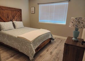 Une chambre à coucher avec un lit, une tête de lit en bois, une fenêtre avec des stores et une table de chevet avec une lampe.