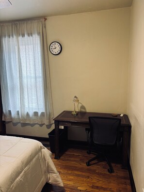 Ein Hotelzimmer mit einem Bett, einem Schreibtisch mit Stuhl, einer Wanduhr und einem Fenster mit durchsichtigen Vorhängen.