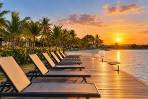 A wooden deck with lounge chairs facing a body of water, palm trees, and a sunset. - Lovely 2-bedroom apartment with AC in vibrant Punta Cana (Punta Cana)
