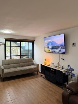 Una sala de estar con un sofá, un televisor montado en la pared, una ventana y una mesita con adornos.