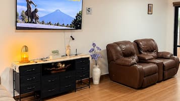Un salon comprenant un téléviseur à écran plat, un canapé, un bureau avec des tiroirs, un vase de fleurs et une cheminée.