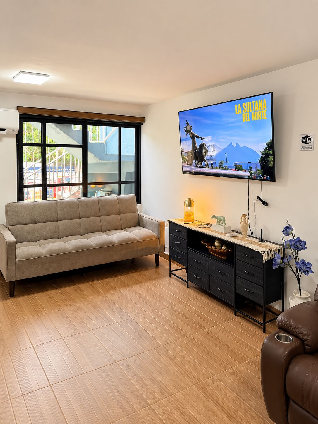 Sala de estar com sofá, televisão na parede, escrivaninha com gavetas e janela com vista para uma montanha.