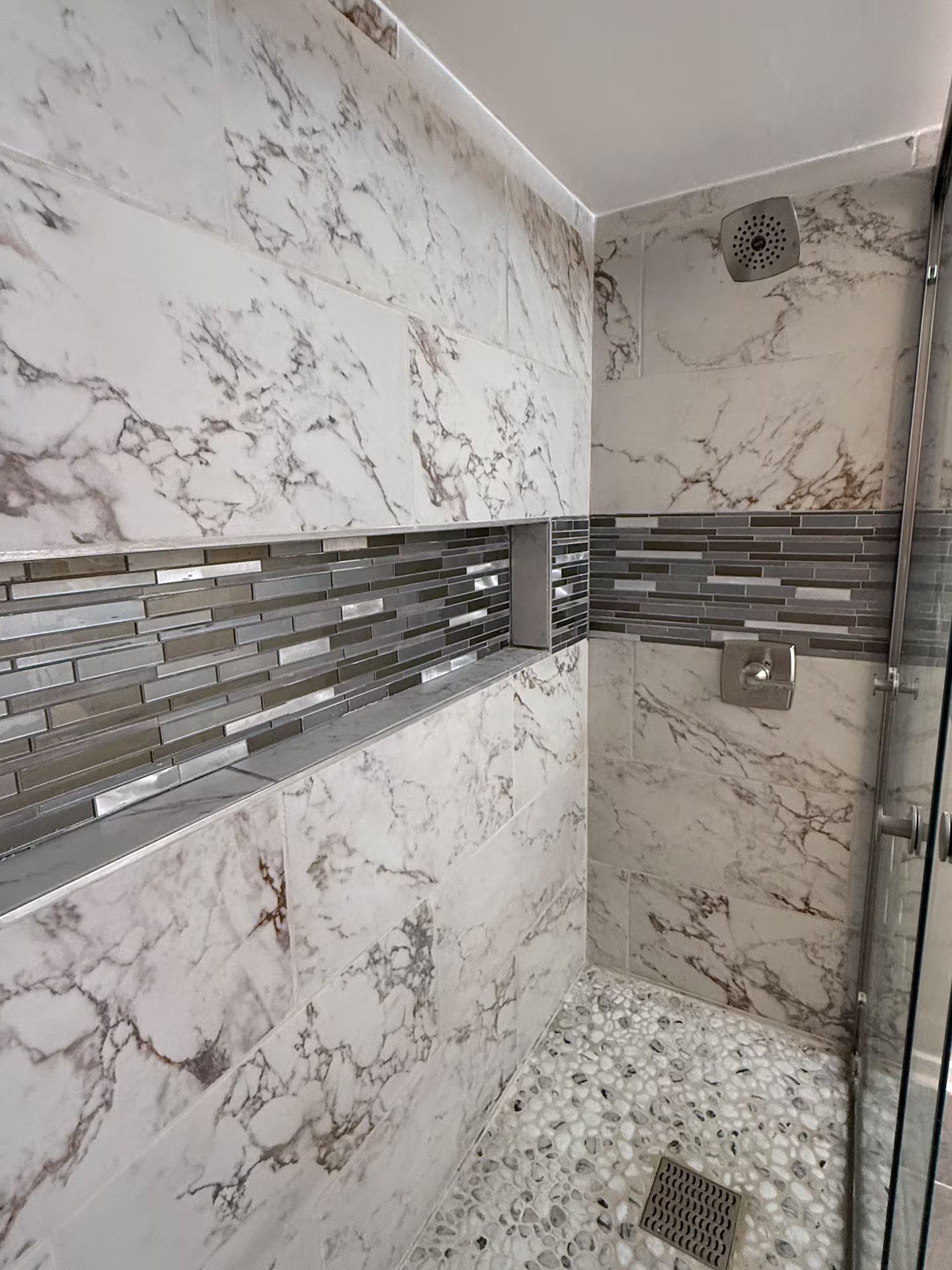Un baño con paredes de mármol, zona de ducha con azulejos y mampara de cristal.