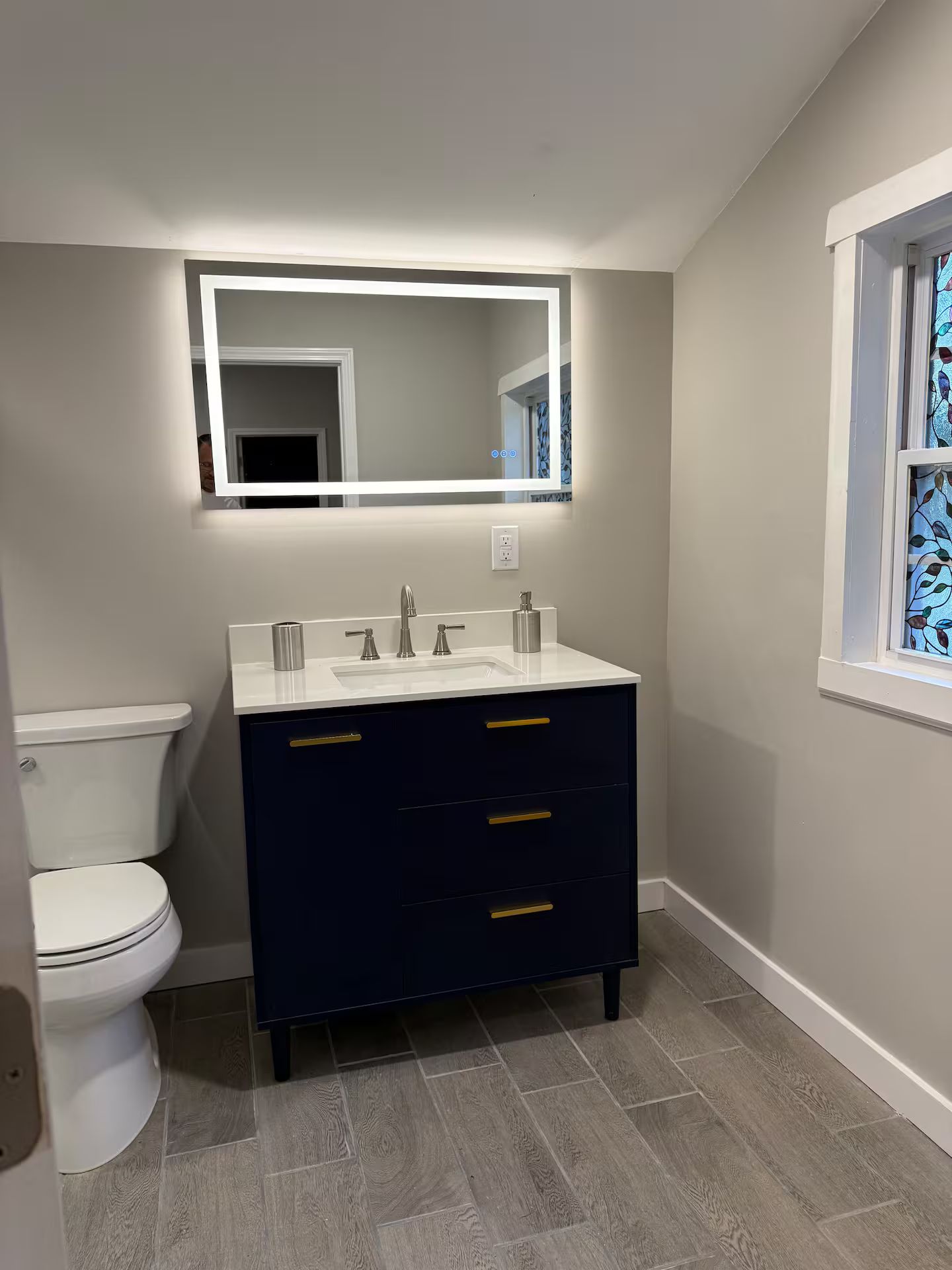 A bathroom with a white toilet, a dark blue vanity with gold handles, and a mirror with integrated lighting.