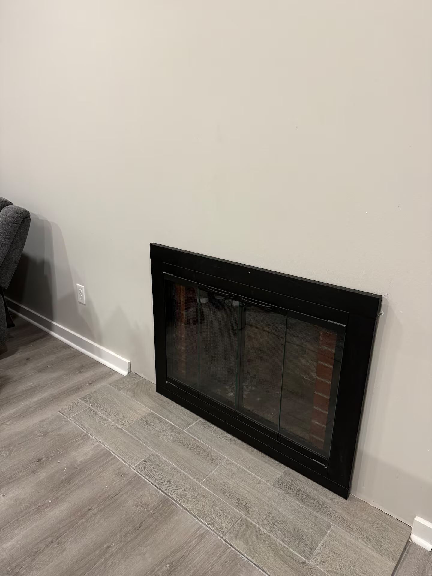 A fireplace with a black frame and glass doors, set against a plain wall with a grey floor.