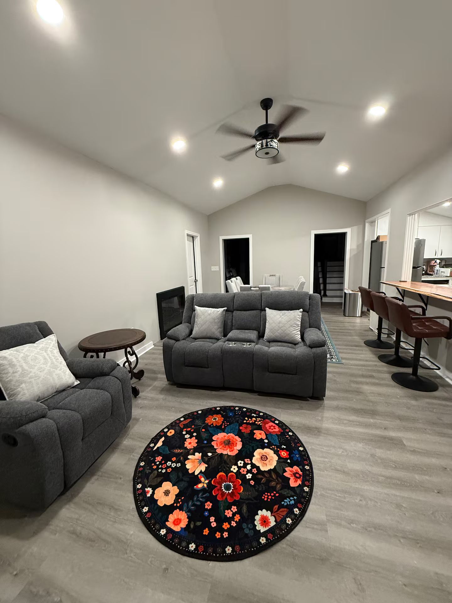 A modern living room with a grey sofa, a round rug with floral patterns, a ceiling fan, and a kitchen area in the background.