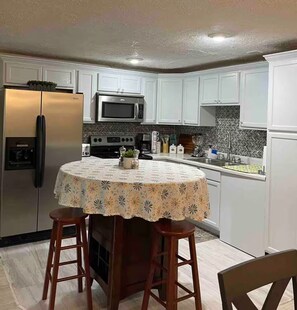 A kitchen with white cabinets, a stainless steel refrigerator, a microwave, and a stove. There is a round table with a floral tablecloth and two wooden stools. - Modern stay-near Fort Knox (Radcliff)