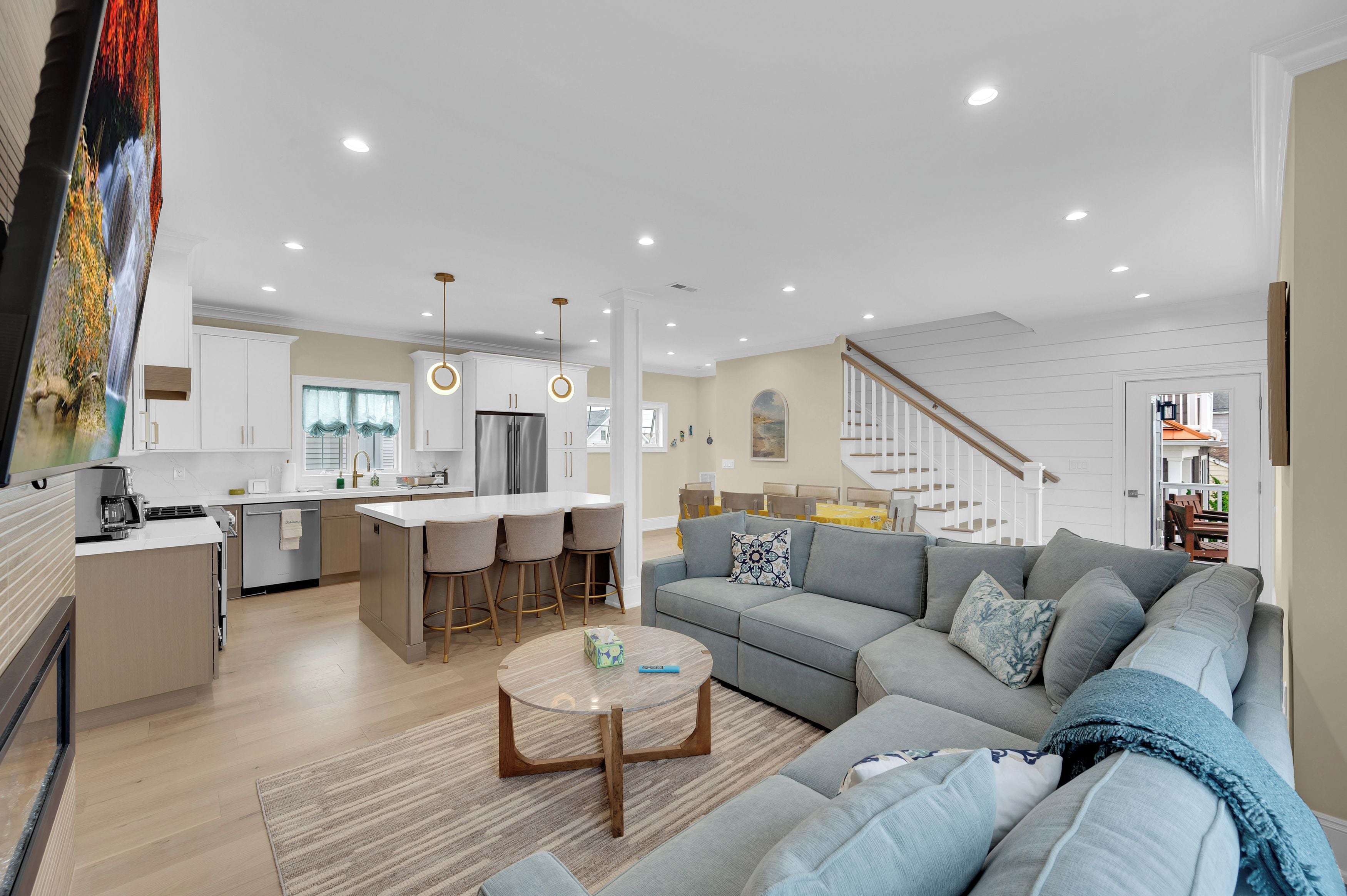 A modern living room with a grey sofa, a coffee table, and a kitchen area with white cabinets and a stainless steel refrigerator.