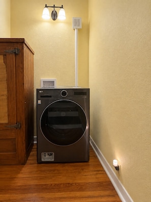 A compact, modern laundry room with a front-loading washer and dryer, a wooden cabinet, and a wall-mounted light fixture. - Downtown Escape #2- New Listing! (Pensacola)